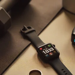 A sleek flat-lay of modern wearable fitness monitors, including a smartwatch, a smart ring, and a fitness tracker band placed on a gym mat with dumbbells and a water bottle. The smartwatch screen displays heart rate and sleep tracking data, while the fitness band shows steps and calories burned. The setup conveys technology, fitness, and lifestyle integration in 2025.