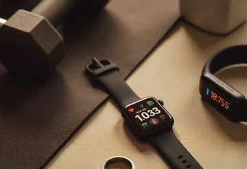 A sleek flat-lay of modern wearable fitness monitors, including a smartwatch, a smart ring, and a fitness tracker band placed on a gym mat with dumbbells and a water bottle. The smartwatch screen displays heart rate and sleep tracking data, while the fitness band shows steps and calories burned. The setup conveys technology, fitness, and lifestyle integration in 2025.